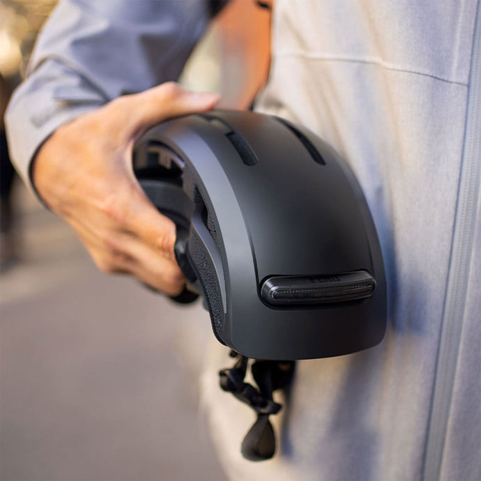 Urban cyclist holding a black folded FEND Super portable helmet outdoors, wearing a gray jacket.