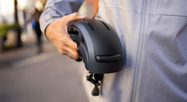 Cyclist holding a black folded FEND Super bike helmet outdoors, wearing a gray jacket.