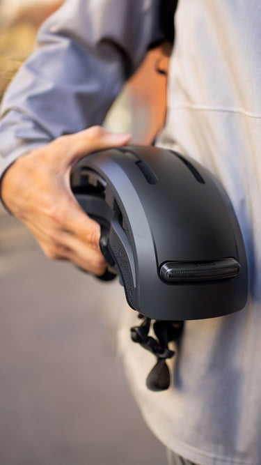 Cyclist holding a black folded FEND Super bike helmet outdoors, wearing a gray jacket.