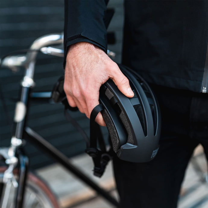 Man holding a black folded FEND One urban commuter bike helmet with a bicycle parked against the wall in the background.