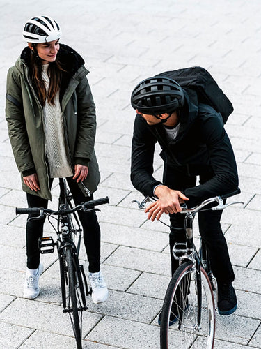 Urban commuters, man and woman, wearing FEND One folding bike helmets, standing with their bicycles on gray city tiles.